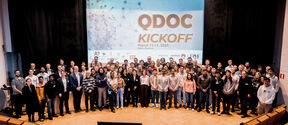 Iso ryhmä ihmisiä seisoo näytön edessä, jossa lukee 'QDOC KICKOFF March 11-12, 2025'.