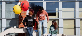 Three students in Aalto hoodies hang out by a brick wall with balloons and metal steps