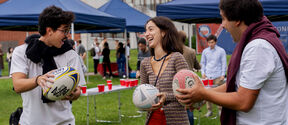Kolme ihmistä ulkona nurmella pitelee rugby-palloja käsissään, taustalla pöytiä ja sinisiä telttoja