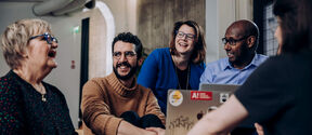 Group of adults sit around a stickered laptop, talking together in a modern room with pale walls.