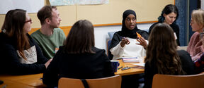 Group of people sit around a wooden table discussing, with large maps pinned on the wall behind them.