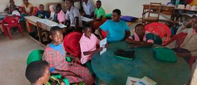 Children sit around tables in a classroom with red and green chairs, reading and talking together.