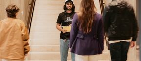 Student in black Aalto University shirt stands on indoor stairs facing three approaching people