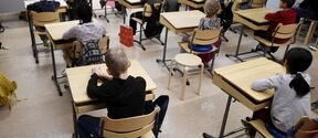 Primary pupils sit spaced at wooden desks in a bright classroom, facing the teacher at the front.