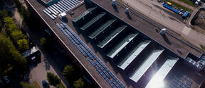 Aerial view of large flat-roof building with glass skylights, solar panels and nearby road with parked vehicles