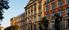 Historic stone and red-brick building with arched entrance, tall windows and people walking along the street. It's TU Darmstadt, Germany.