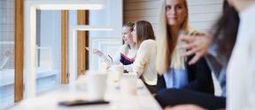 People sit by tall windows at a bright white counter with lamps and coffee cups, talking together