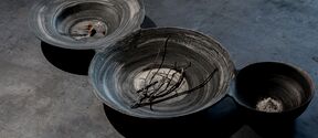 Three linked grey ceramic bowls on a dark table, holding twigs, ash and small debris