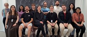 Group of people posing indoors in two rows, some seated on stools, chalkboard behind