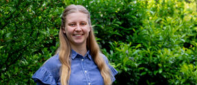 Person with long hair in a blue shirt standing in front of dense green bushes