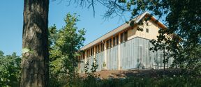 Modern light wood and metal building on a slope, surrounded by tall green trees under blue sky