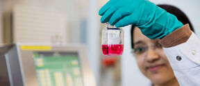 Lab worker holds clear vial of pink liquid near a machine with a green-screen display