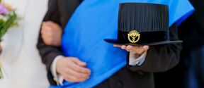 Person in formal suit with blue sash holding a black doctoral hat with a gold emblem