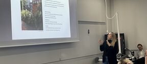 Maiju Suomi giving a lecture in a grey classroom, gesturing towards a large slide with text and a garden photo.