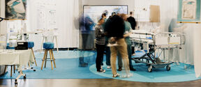 Blurred people around a hospital bed in a bright clinic room with blue floor and medical equipment