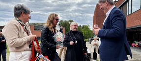 Group chatting outdoors by red brick buildings; woman holds gift flowers, man in blue blazer with drink