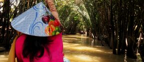 Person in a fruit-print conical hat paddling a small boat through a narrow palm-lined river. Photo by Liisa Perälä, Mekong Delta, Vietnam, 2026