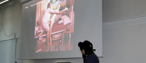 Person in dark clothes presents a colourful bedroom photo on a large white projection screen in a classroom.