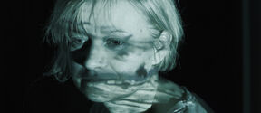 Person with light hair in a dark room, face obscured, bluish light projected on white shirt