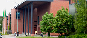 Modern red-brick building with tall black pillars, trees in front and people walking on the pavement