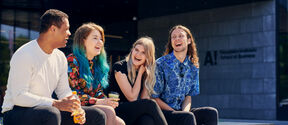 Four people sit chatting with drinks on a campus bench outside a modern university building.