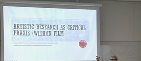 Lecture on artistic film research, presenter gesturing beside large projected slide in bright classroom.