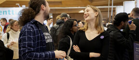 Two people talk at a busy indoor event, standing among a crowd under warm wooden ceiling.