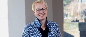 Smiling person with short blond hair and glasses in a zigzag blue-white jacket, standing indoors by a window