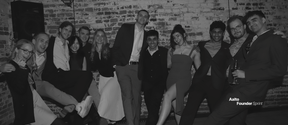 Group of smartly dressed people posing together at a brick‑walled indoor event, some holding drinks.