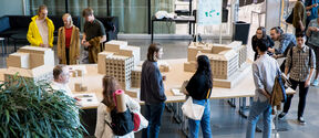 People in casual clothes discuss beige architectural models on tables in a bright hall