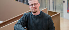 Man with glasses and short beard in dark jumper, leaning on railing in bright modern office