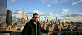 Man in dark coat on rooftop terrace with New York skyline and cloudy blue sky behind