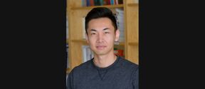 Person with blurred face in a dark grey top standing indoors in front of wooden bookshelves.
