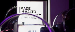 Glowing Aalto University sign in a dark space, seen through clear round chairs lit with purple light