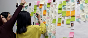 Three people add notes to a white wall filled with colourful sticky notes and small printed photos
