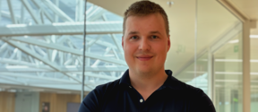 Person with blurred face in dark polo shirt, arms crossed, standing in bright modern glass office