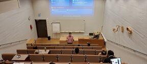 Man giving lecture on textile chemistry in tiered auditorium, students seated with laptops and notes