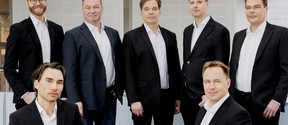 The PulseOn team posing for the camera. 7 men in suits, 5 standing and 2 sitting on the sides