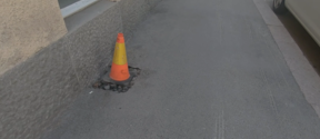 Orange and yellow traffic cone marking a small hole in a grey pavement by a building wall