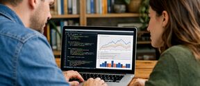 Two people at a wooden table review code and charts on a laptop, with bookshelves and plants behind them