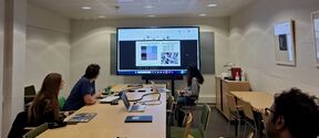 Four people in a meeting room watch a large screen showing charts and photos, with laptops on a long table.