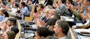 Large, diverse audience in a lecture hall applauding, seated in curved rows with laptops and notebooks