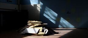 Sunlit pile of wooden and white boards on a gym floor in a large blue-walled hall