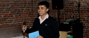 Smiling young man speaking into a microphone, holding blue papers, standing by brick wall indoors