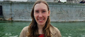 Smiling young woman in tan coat by green fountain pool with stone sculptures in the background
