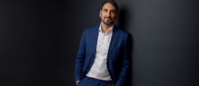 Smiling man in a blue suit and white shirt leaning against a dark wall