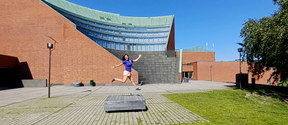 Person in blue shirt jumps joyfully on a paved campus yard before a curved brick and glass building