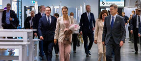 Smiling woman in a pale suit holding flowers walks with suited group through a modern indoor venue