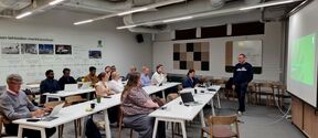 People seated at long white tables with laptops as a man presents slides in a modern classroom.