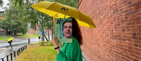 Person in green holds magazine over face under yellow Aalto umbrella by wet brick wall and trees